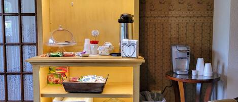 A small, well-lit room with a wooden cabinet containing a coffee maker, cups, and snacks. A bed and a chair are visible in the background.