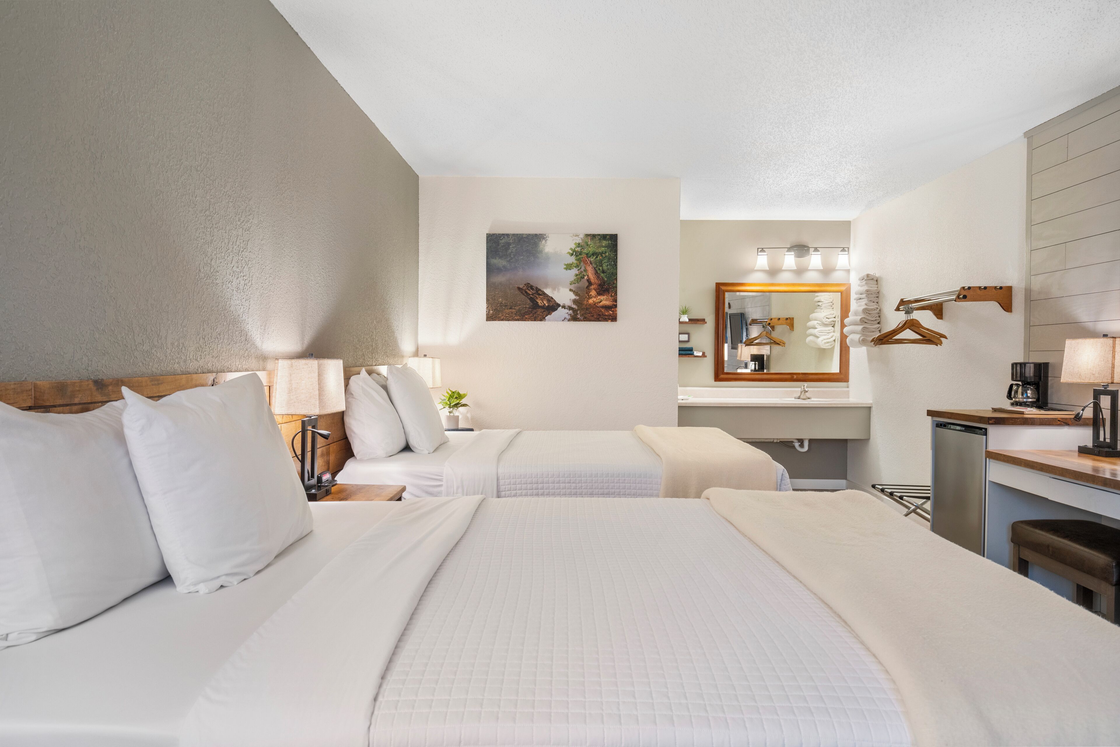 A hotel room with two beds, a bathroom with a mirror and towel rack, and a small kitchenette area.