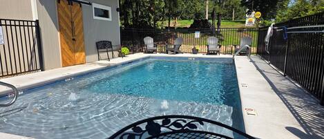 A swimming pool with a wooden deck, lounge chairs, and a small building with a door.
