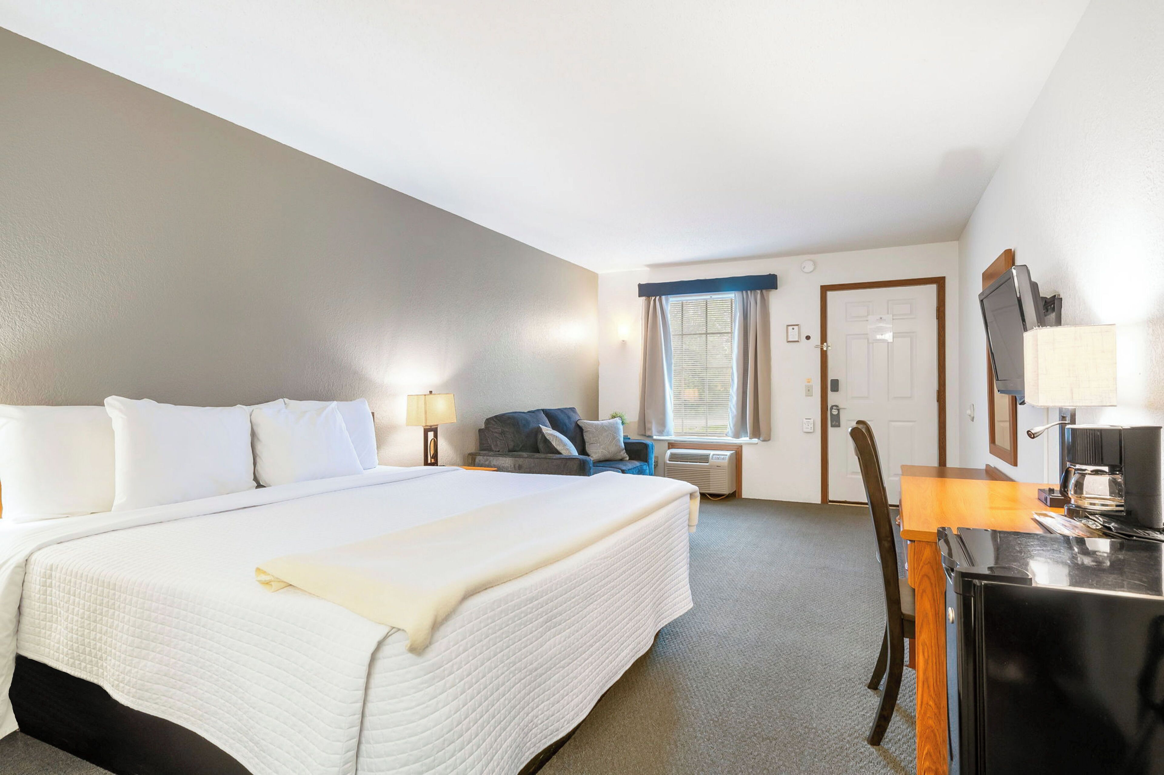 Ein Hotelzimmer mit einem großen Bett, einem Schreibtisch mit Computer, einem Sessel, einem Fenster mit Vorhängen und einer Tür.