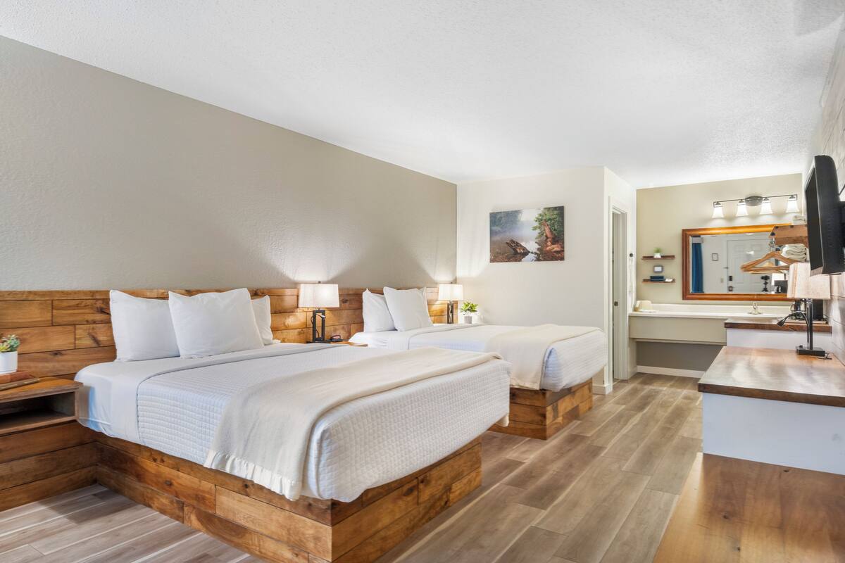 A hotel room with two beds, a wooden headboard, bedside lamps, a small plant, and a bathroom with a mirror and sink.