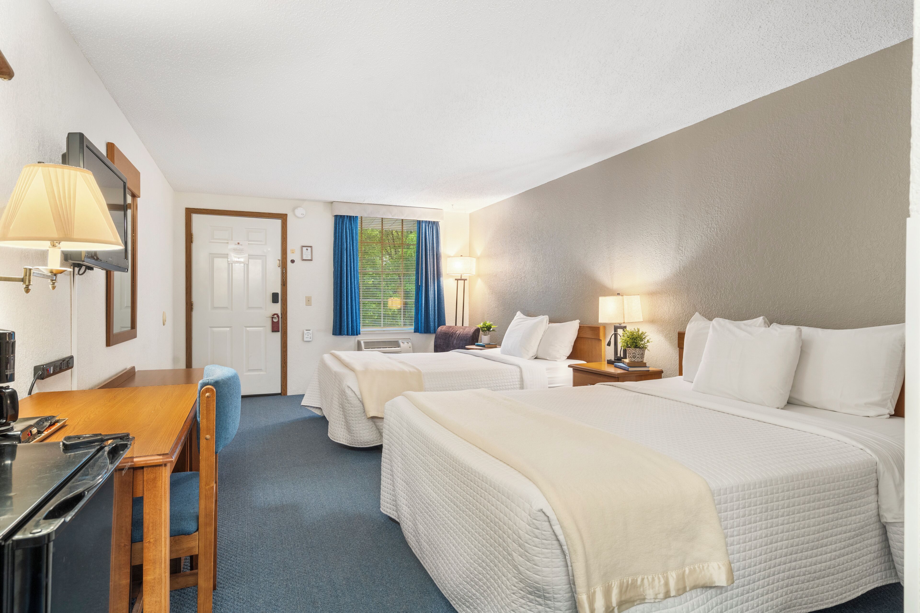 A hotel room with two beds, a desk with a chair, a TV, and a window with blue curtains.