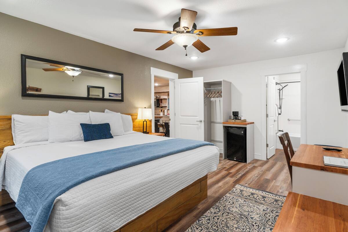 A bedroom with a bed, bedside table, lamp, ceiling fan, and a view into the bathroom.