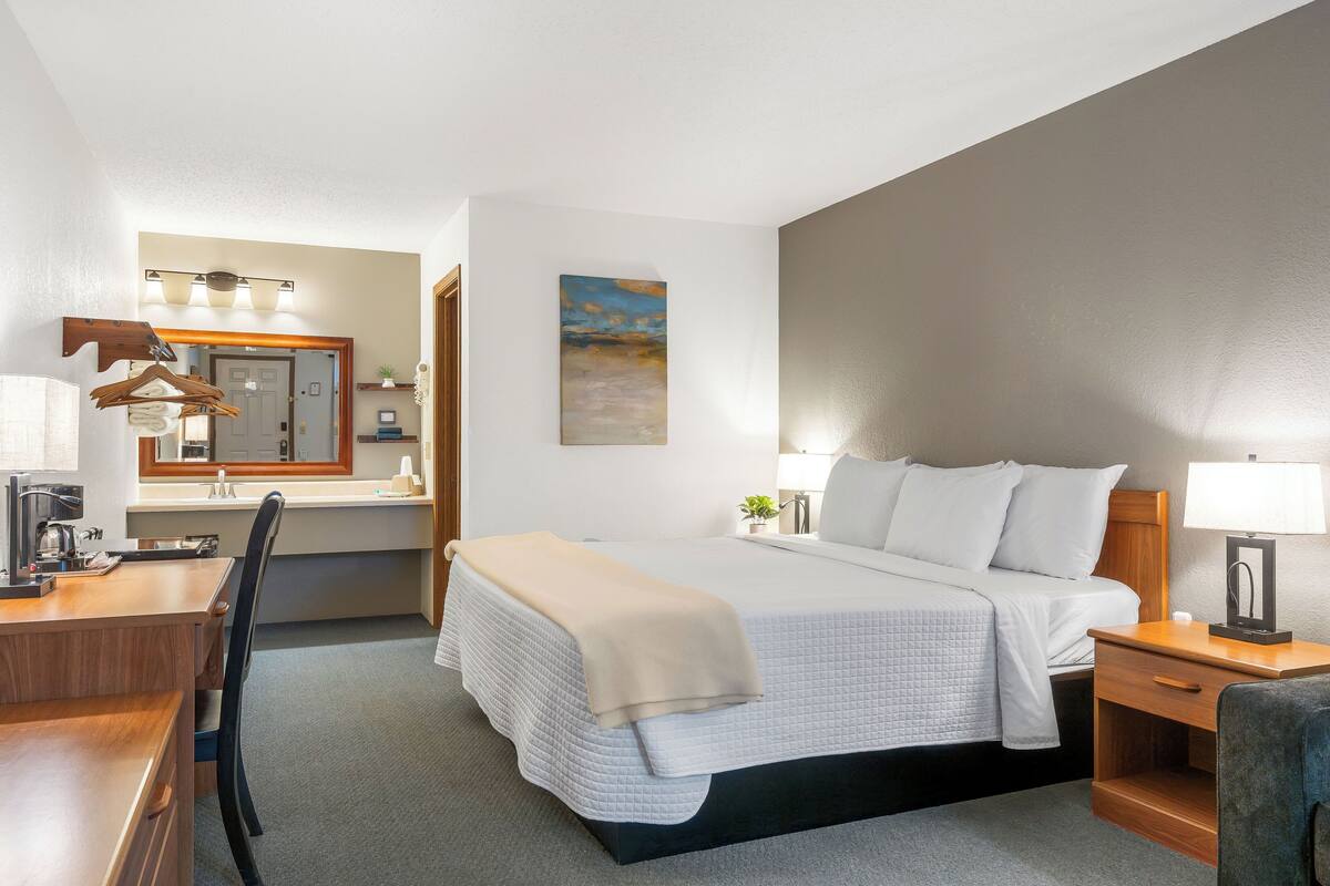 A hotel room with a bed, desk, chair, and a small table with a lamp.