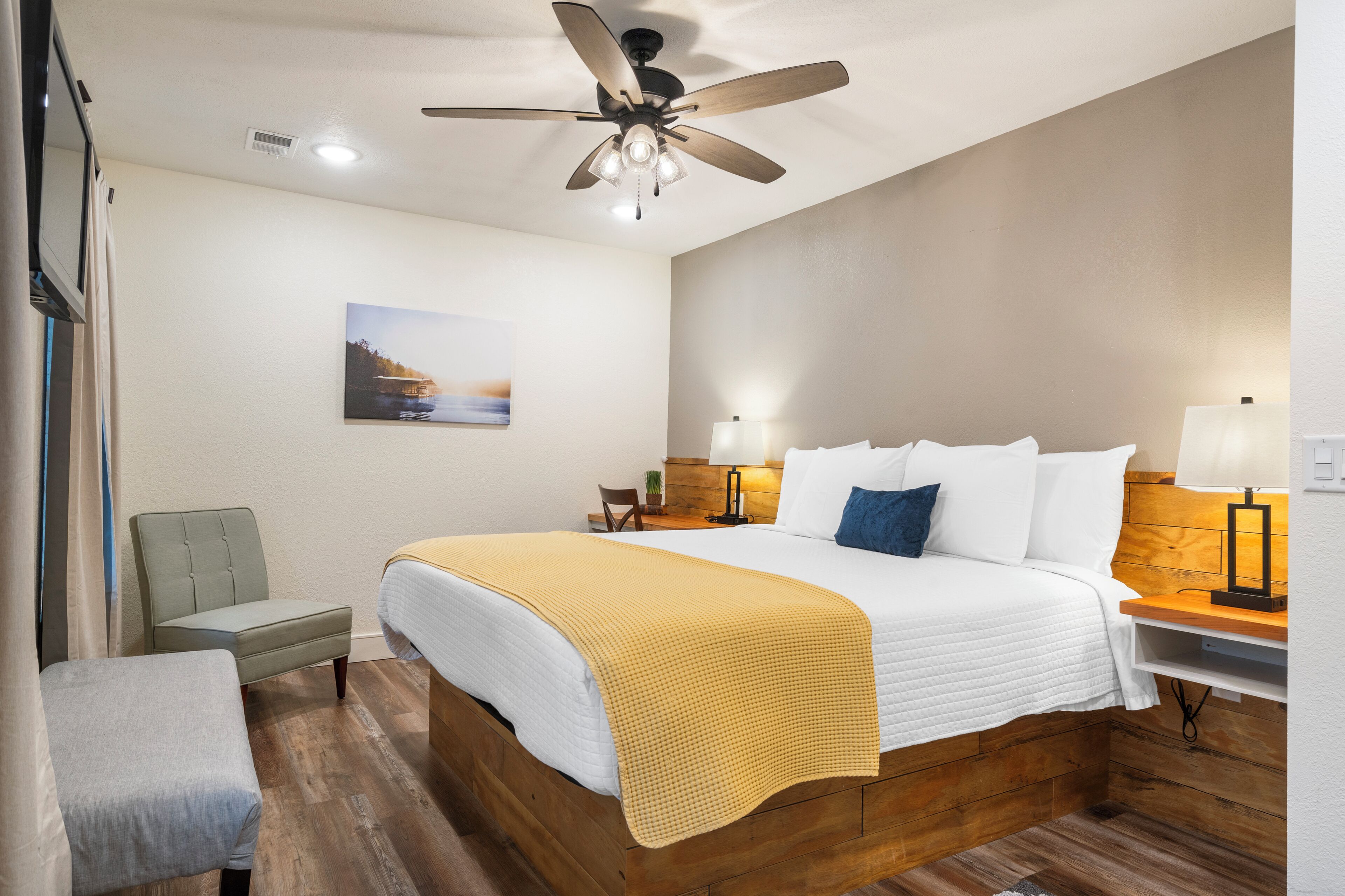 A hotel room with a wooden bed, a ceiling fan, a bedside table with a lamp, a chair, and a painting on the wall.