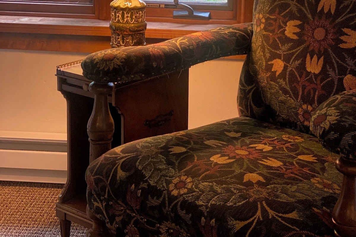 A room with a window, a lamp on a small table, and a floral-patterned chair.