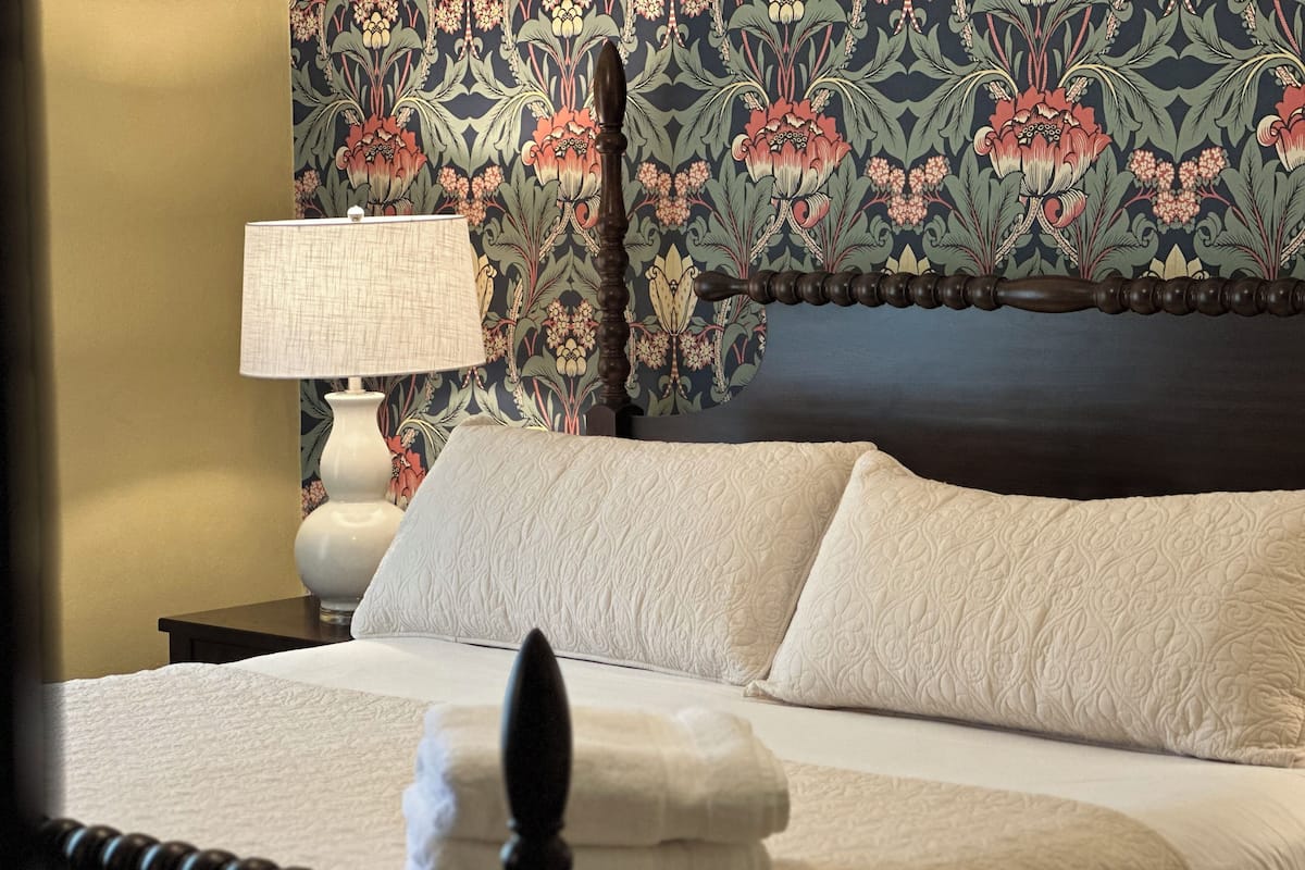 A neatly made bed with a white quilt and two pillows, a bedside table with a lamp, and a floral wallpaper pattern.