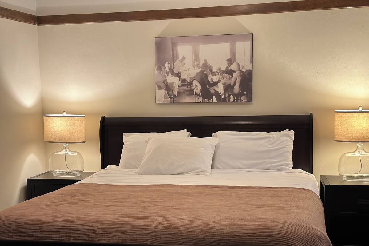 A hotel room with a large bed, two bedside tables with lamps, a framed picture on the wall, and a brown headboard.
