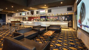 Um saguão de hotel com assentos de couro, área de bar e uma tela grande exibindo um jogo.