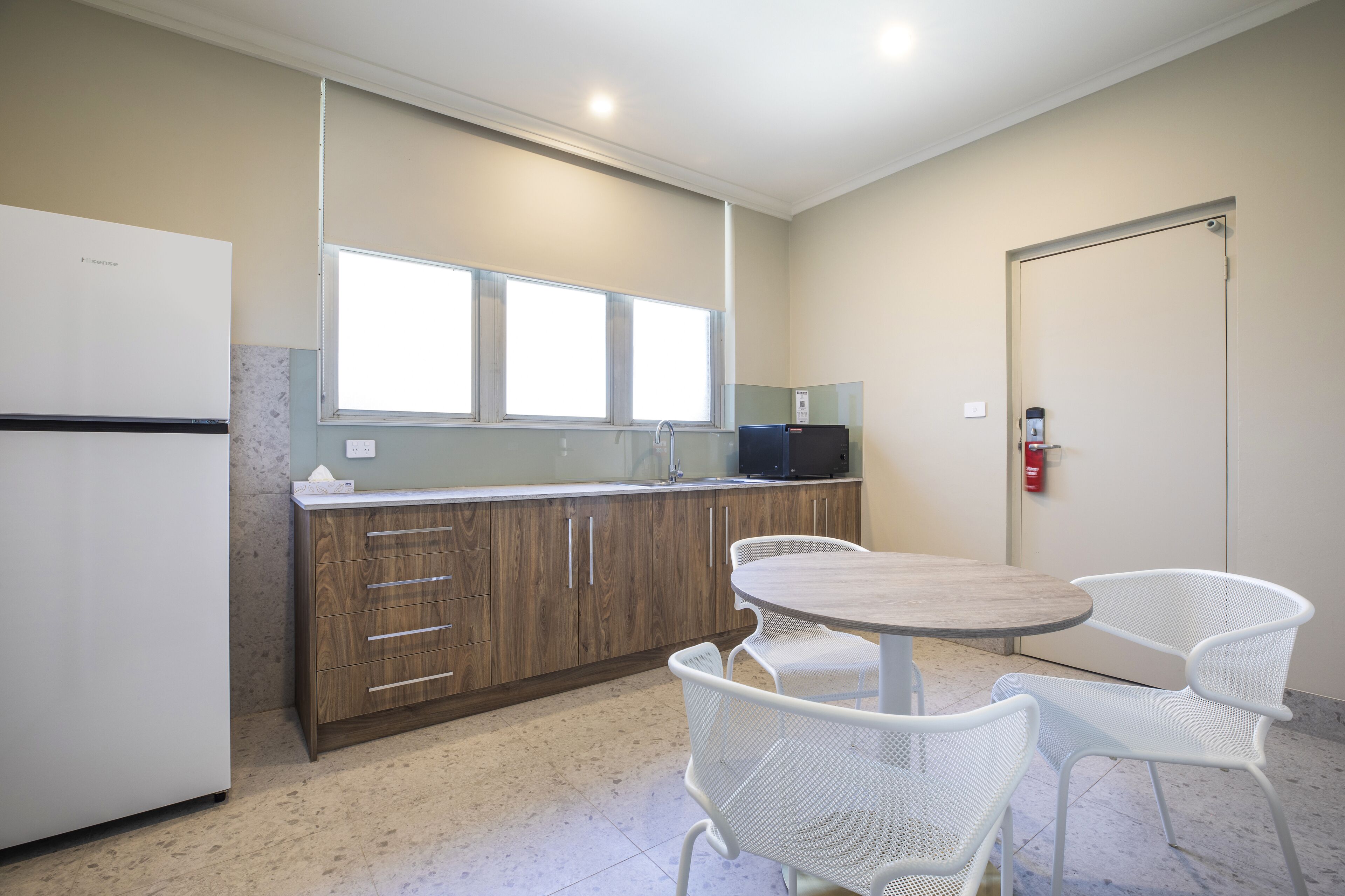 Una cocina compacta con refrigerador, armarios de madera, un lavamanos y una ventana sobre la encimera. Hay una mesa redonda con dos sillas blancas y un extintor de incendios montado en la pared.