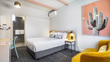 A hotel room with a bed, a desk, a chair, a wall-mounted air conditioner, and a cactus artwork.