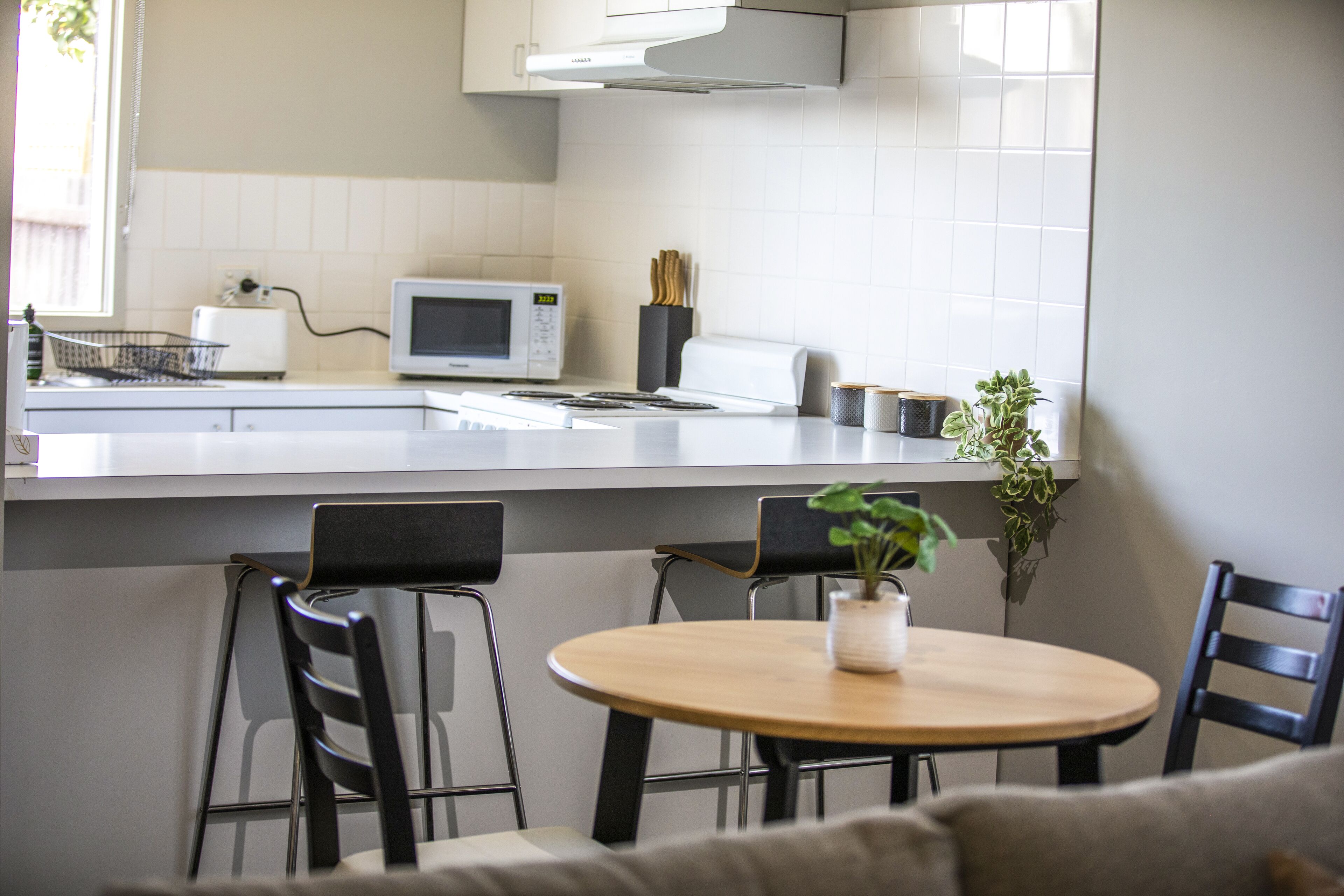 A modern kitchen with white cabinets, a microwave, and a sink. There is a dining table with chairs and a potted plant on the counter.
