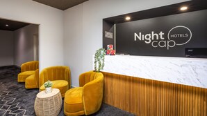 Uma área de recepção com balcão de mármore, uma placa com a inscrição "Nightcap Hotels" e duas poltronas amarelas com uma pequena mesa e uma planta entre elas.