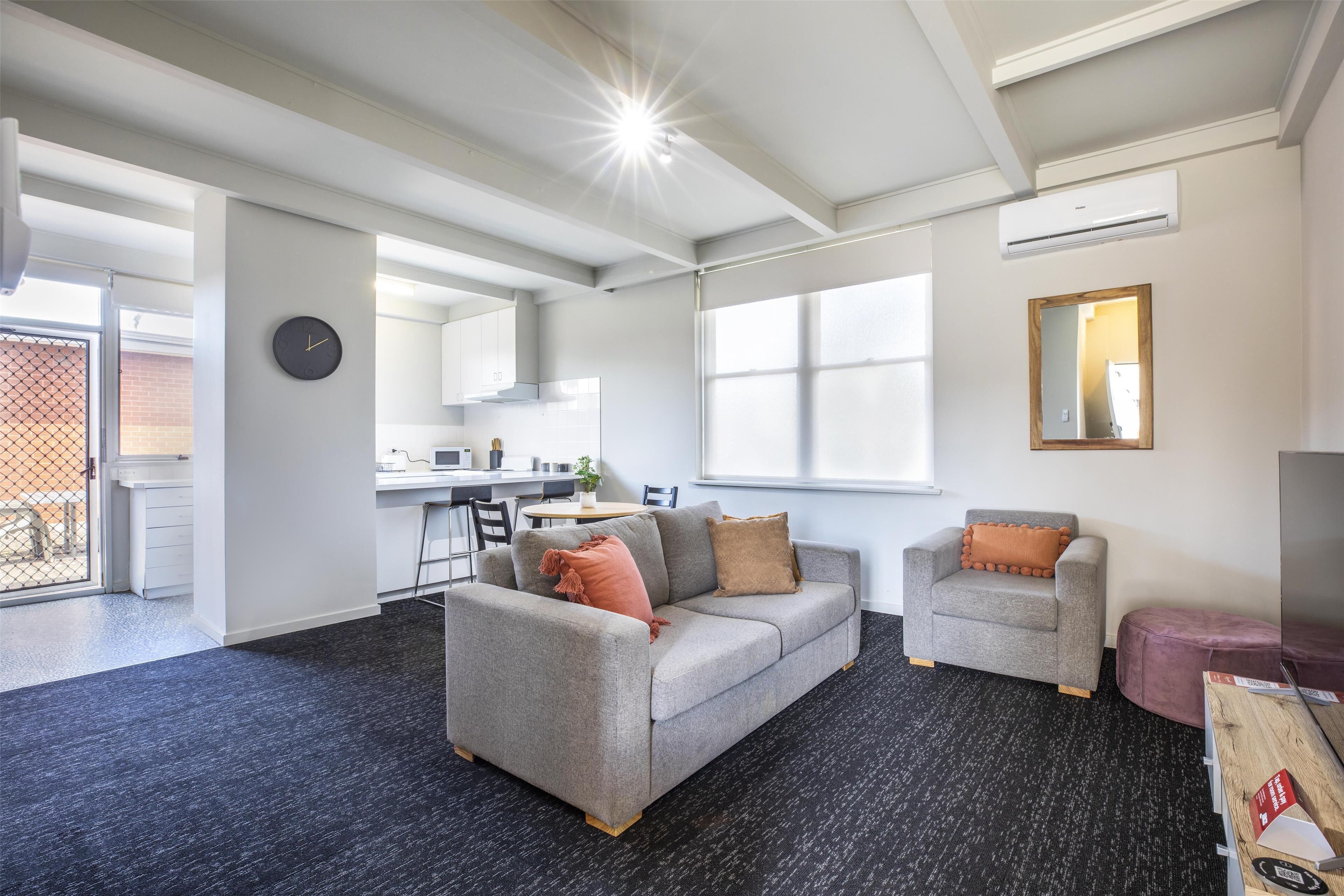 Una sala de estar moderna con un sofá gris, un sillón gris más pequeño y una mesa de centro de madera. Se ve un ventanal que proporciona luz natural, una zona de cocina al fondo y una puerta que da a un espacio exterior.