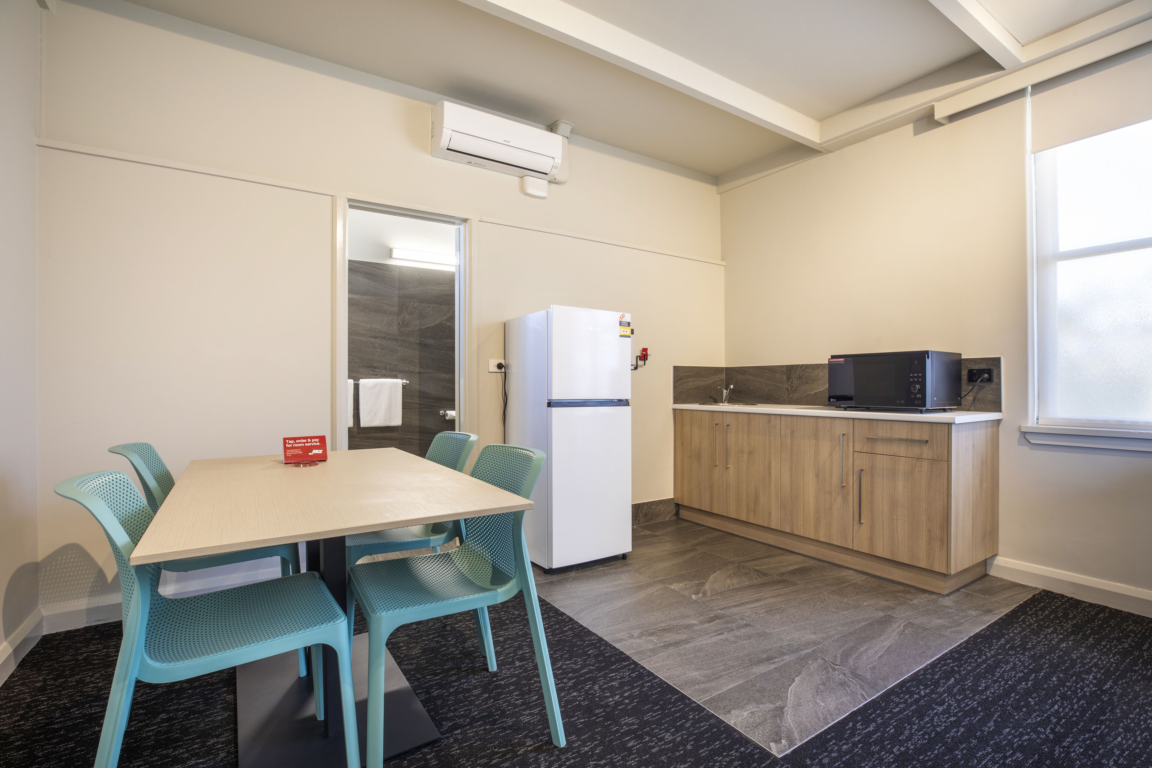 Cocina compacta con refrigerador, microondas y armarios de madera. Hay una mesa de comedor con sillas y una pequeña alfombra en el piso.