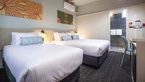 Quarto de hotel com duas camas, uma escrivaninha e um espaço para chuveiro.