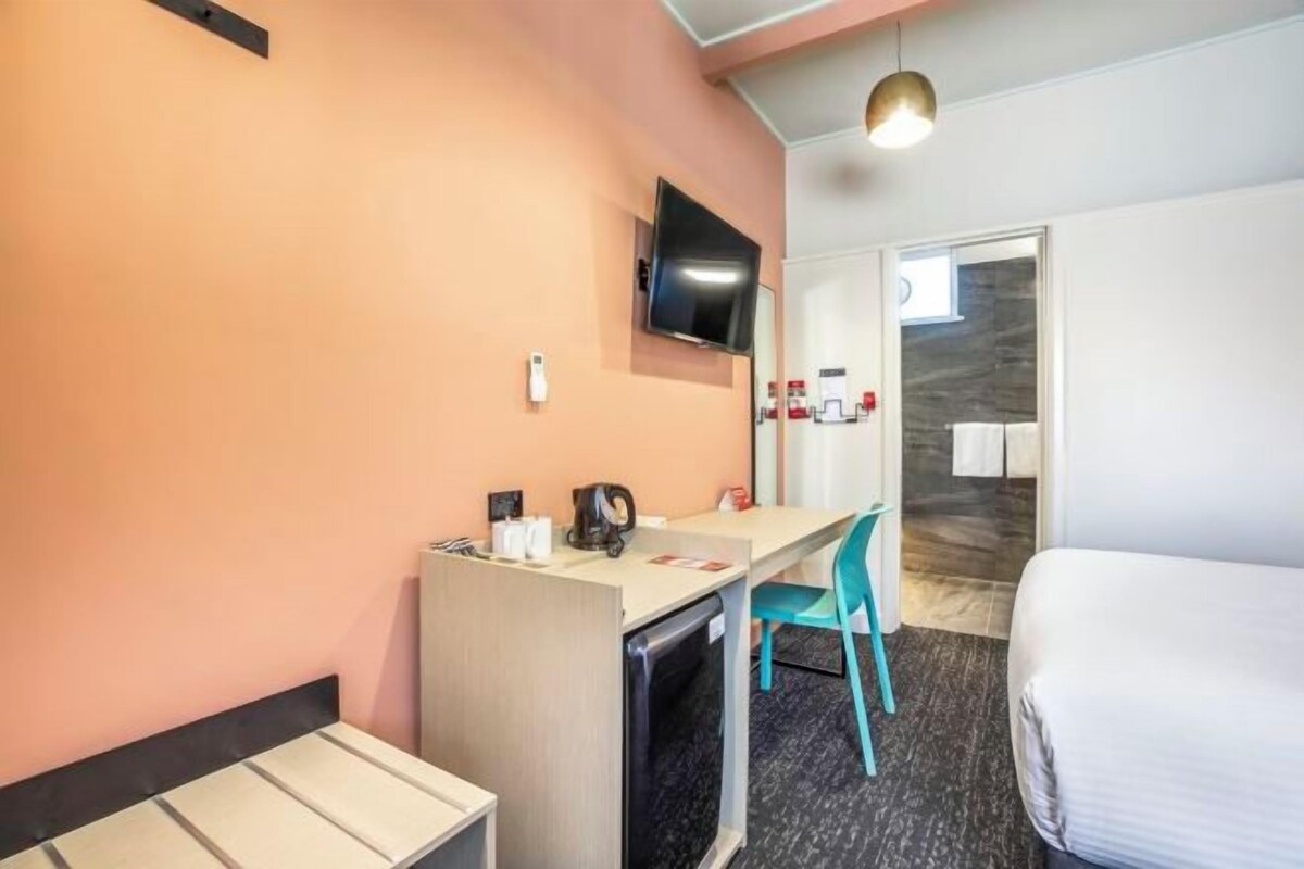 A hotel room with a bed, a desk with a chair, a television mounted on the wall, and a small kitchenette area.