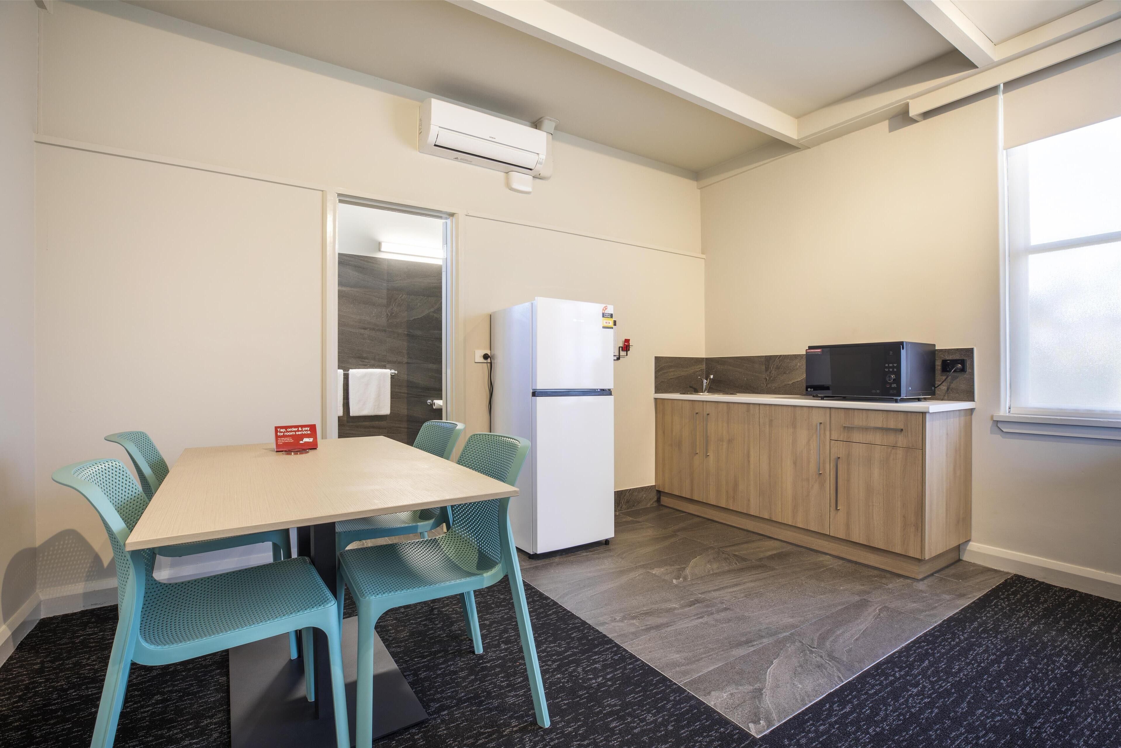 A compact kitchen with a refrigerator, microwave, and a small countertop. There is a dining table with chairs and a window providing natural light.