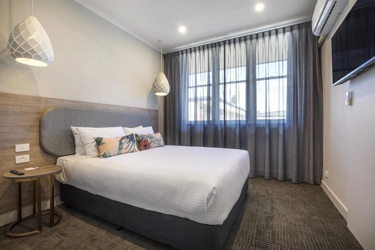 A hotel room with a large bed, a bedside table with a remote control, and a modern pendant light.