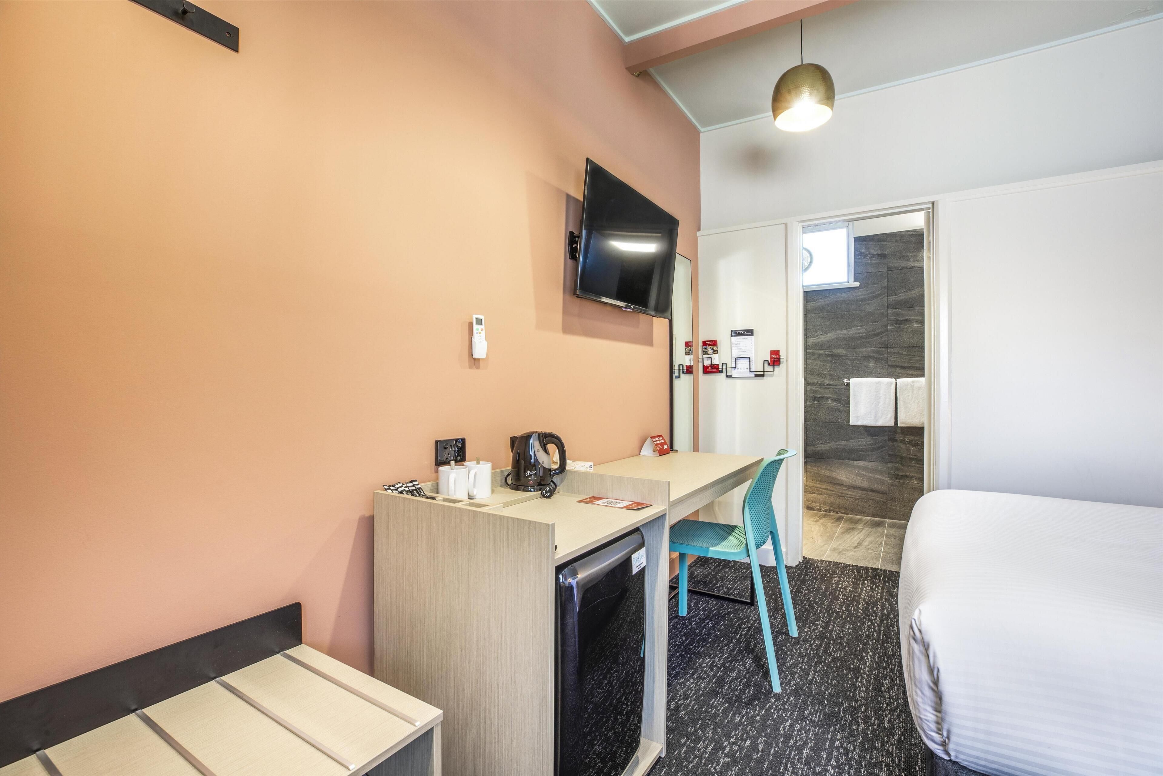 A hotel room with a bed, a desk with a chair, a television mounted on the wall, and a small refrigerator.