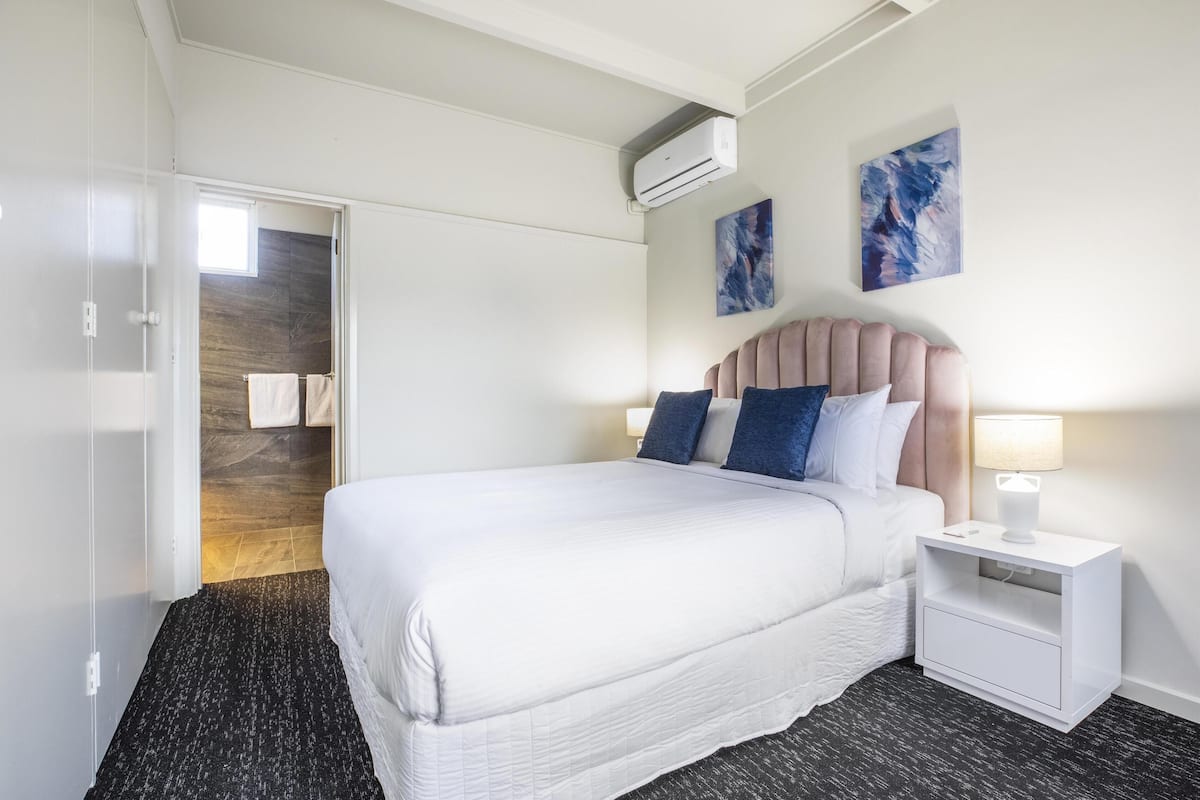 A hotel room with a bed, a nightstand, an air conditioning unit, and two abstract paintings on the wall.