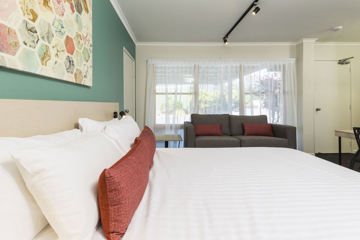 Quarto de hotel com uma cama grande, um sofá, uma escrivaninha e uma janela com cortinas leves.