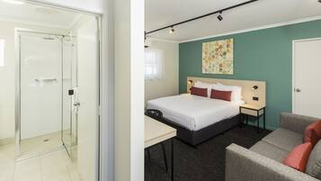Une chambre d’hôtel avec une douche en verre, un lit avec du linge de maison blanc, un canapé gris et une petite table.
