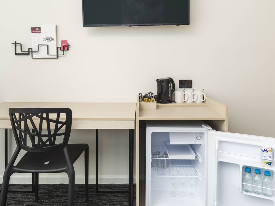 A hotel room with a desk, a chair, a flat-screen TV mounted on the wall, and a mini-fridge with milk bottles.