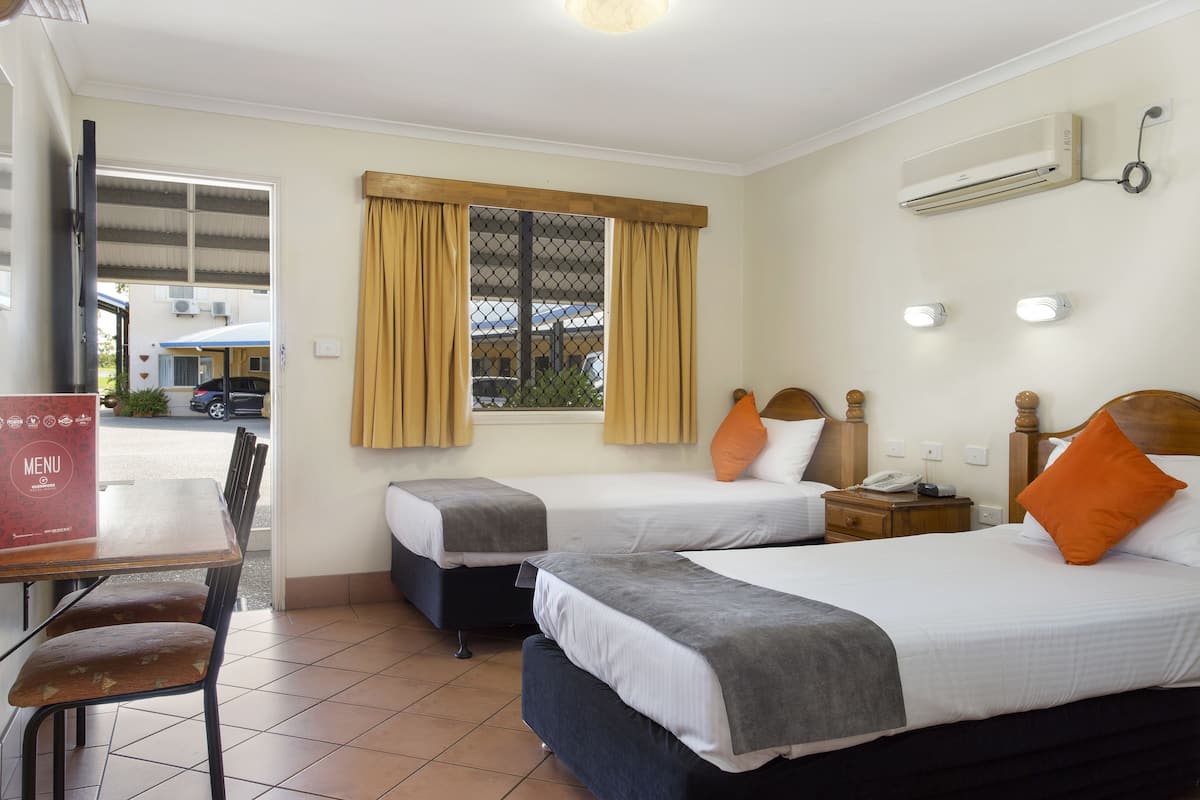 A hotel room with two beds, a dining table, chairs, a window with curtains, an air conditioning unit, and a wall-mounted phone.
