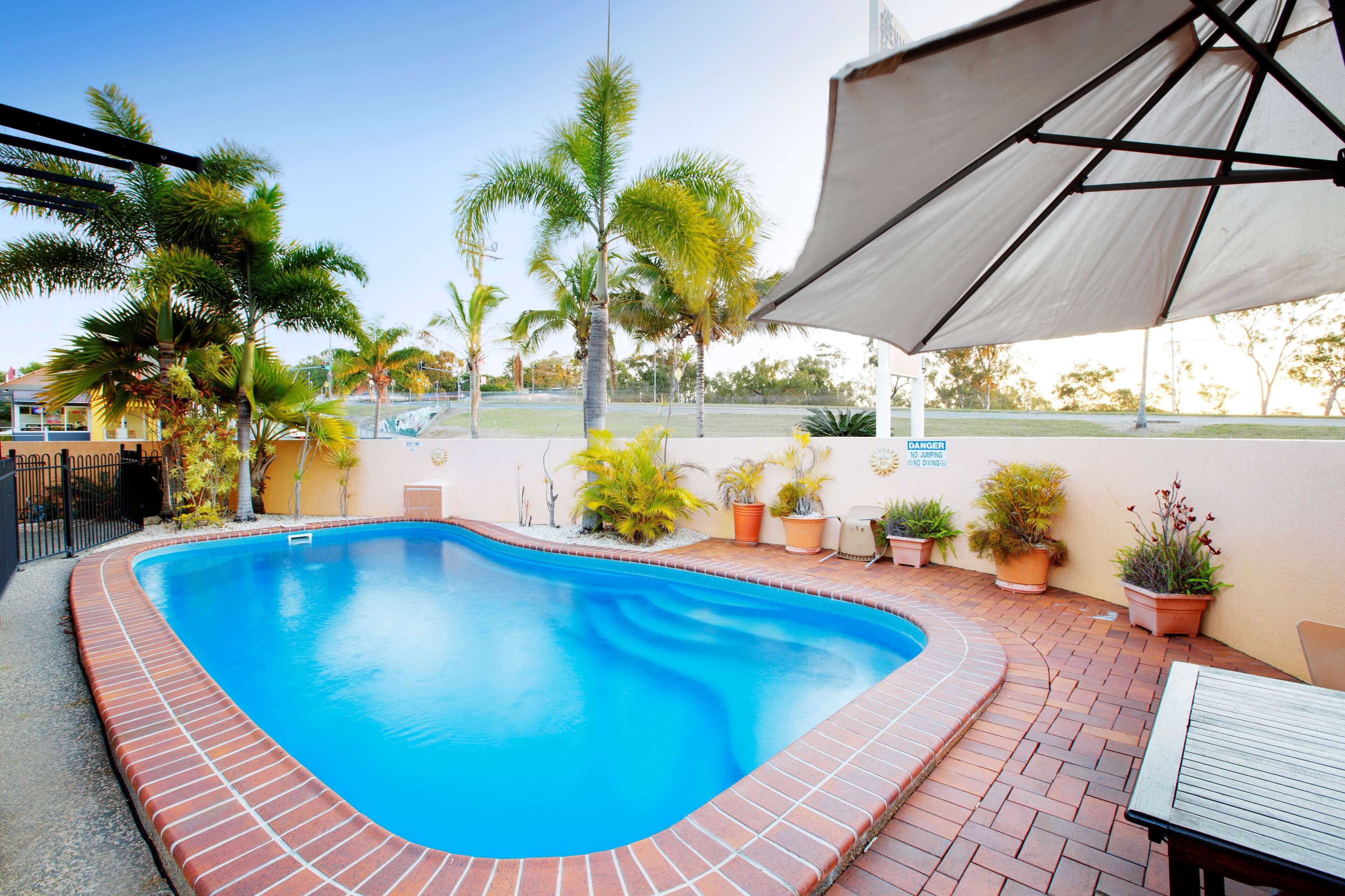 Piscina com borda de tijolos, rodeada por plantas em vasos e uma área coberta com mesa.