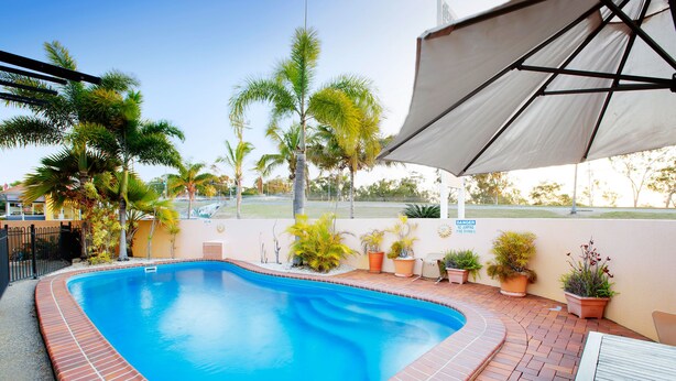 A swimming pool with a brick border, surrounded by potted plants and a covered area with a table and chairs. - Glenmore Tavern (Norman Gardens)