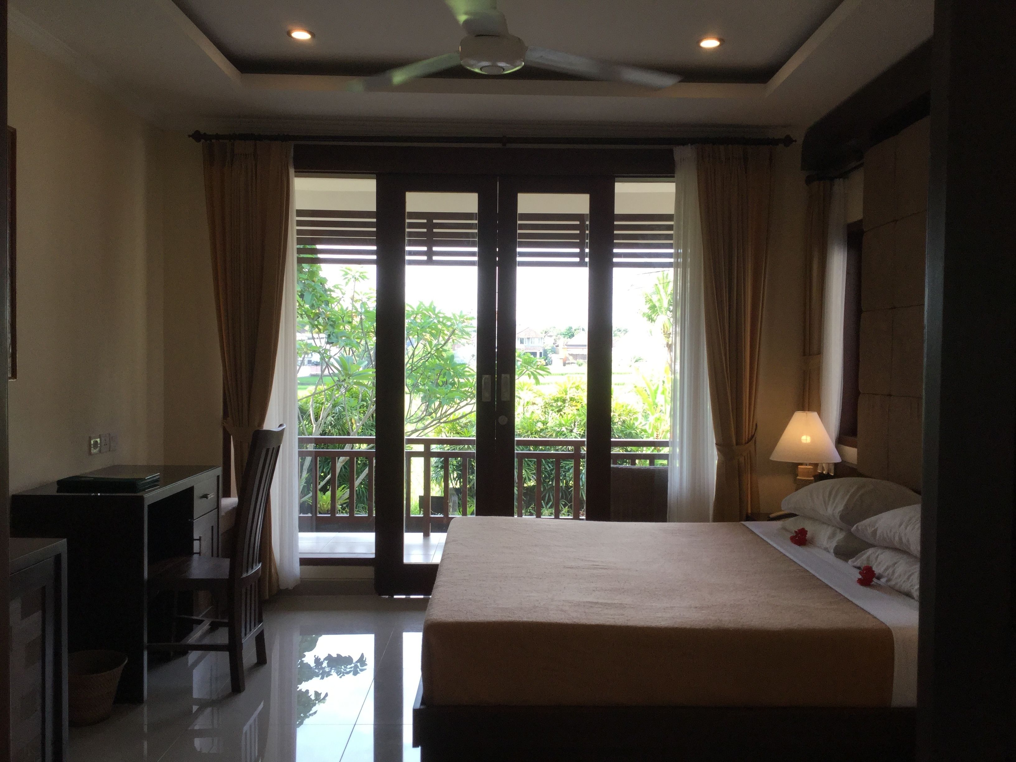 A hotel room with a bed, a desk, a chair, a lamp, and a view of greenery outside.