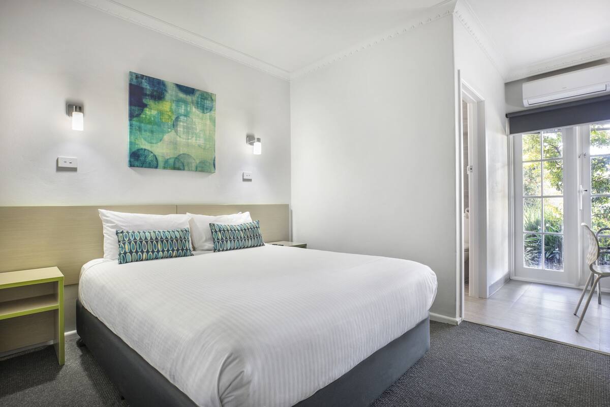 A hotel room with a large bed, a nightstand, a wall-mounted light, and a door leading to a balcony.