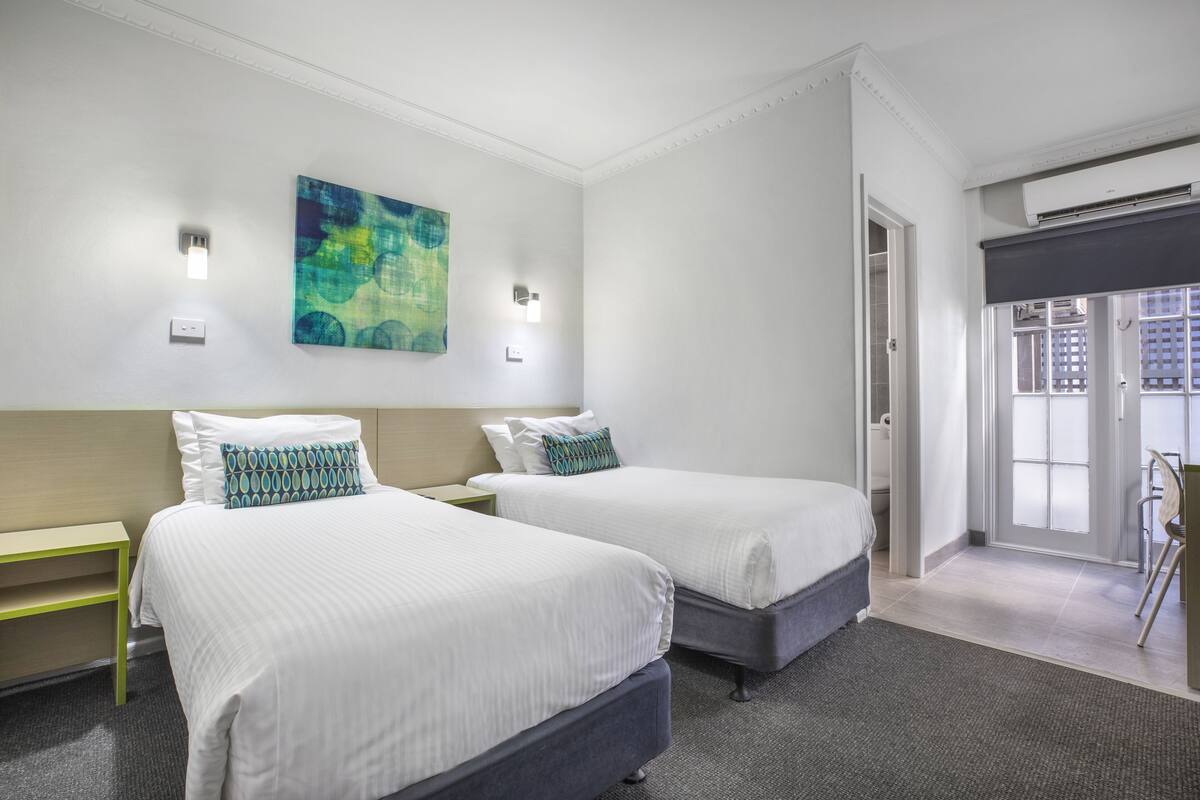 A hotel room with two beds, a painting on the wall, and a view through the door.
