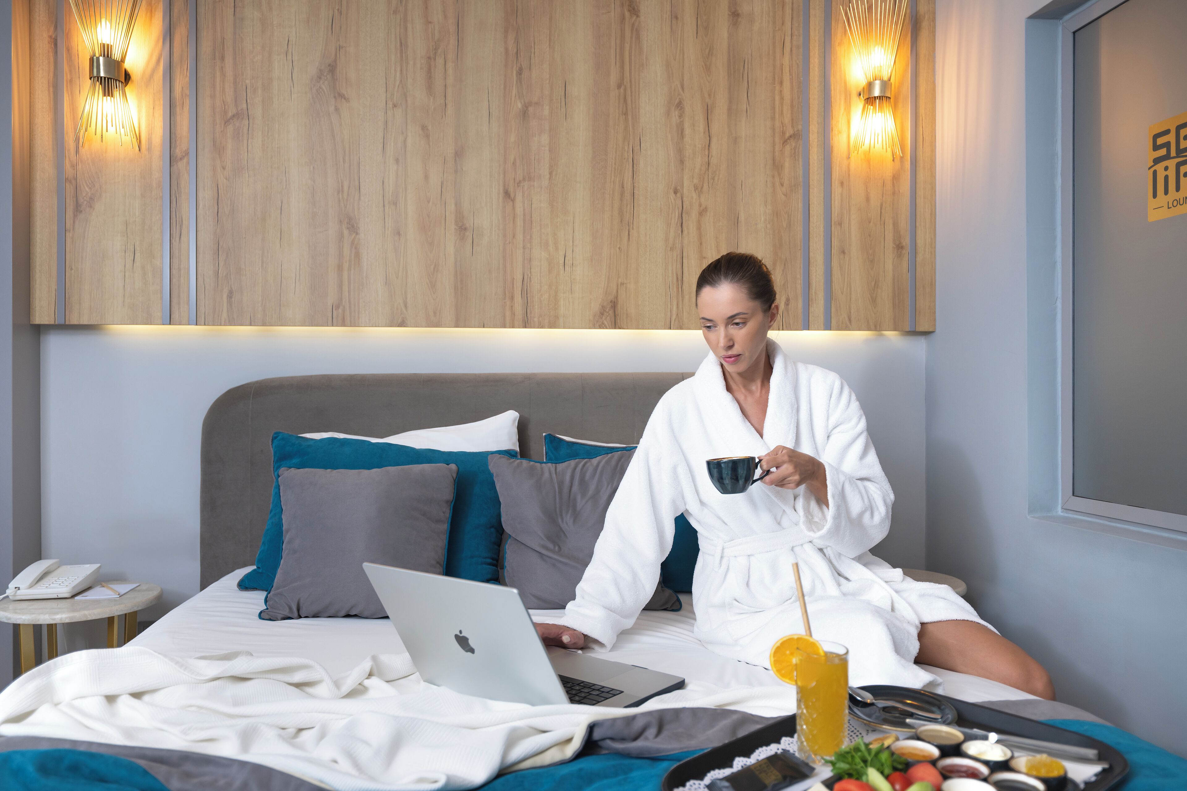 A person in a white bathrobe sitting on a bed with a laptop, a cup, and a tray of food.