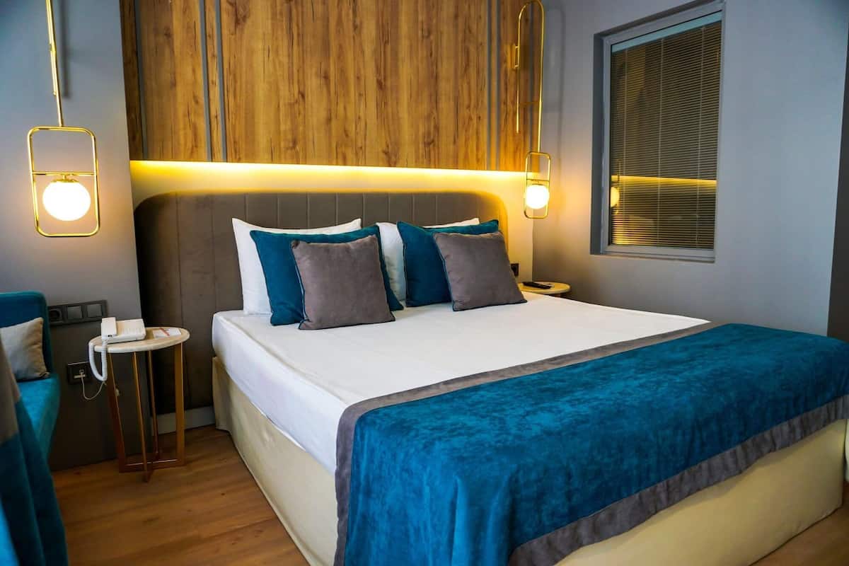 A hotel room with a bed, bedside tables, a wooden headboard, a window with blinds, and a wall-mounted light fixture.