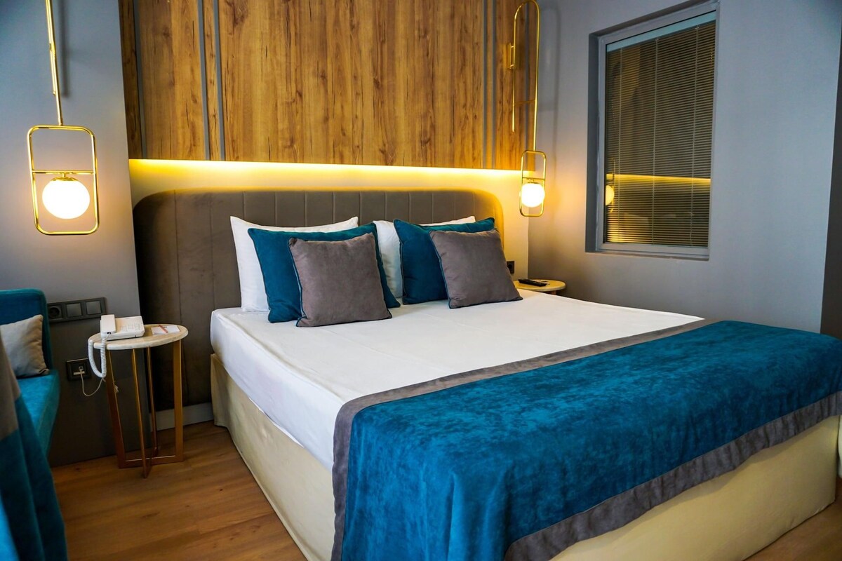 A hotel room with a bed, bedside tables, a wooden headboard, a window with blinds, and a wall-mounted light fixture.