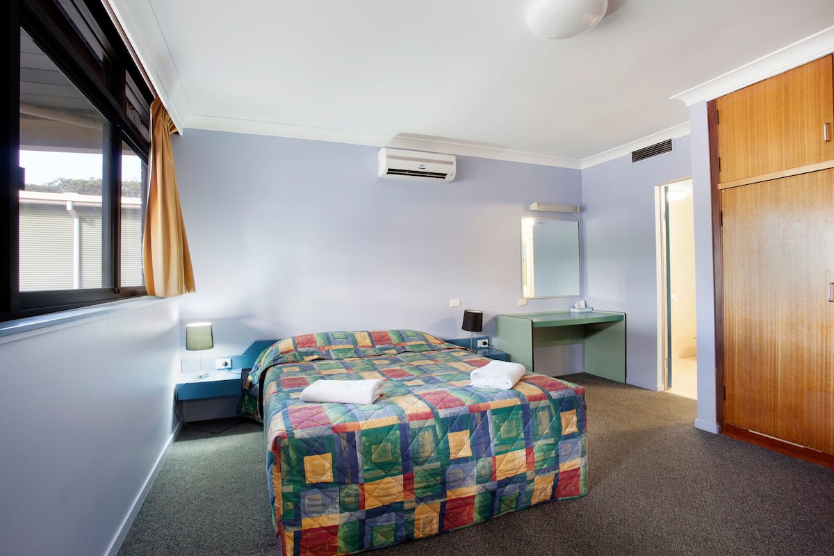 A hotel room with a bed, a desk, a mirror, and an air conditioning unit.