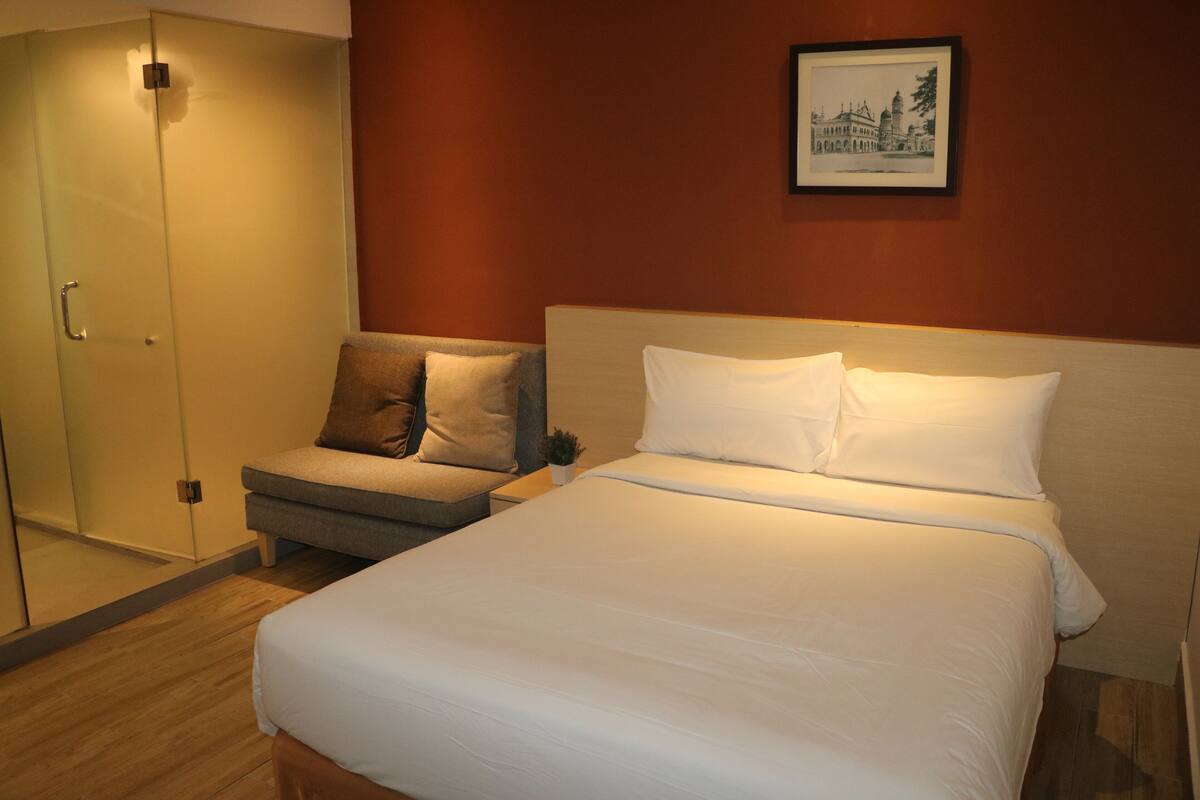 Habitación de hotel con cama, sofá, una mesita y armario con puerta de vidrio.