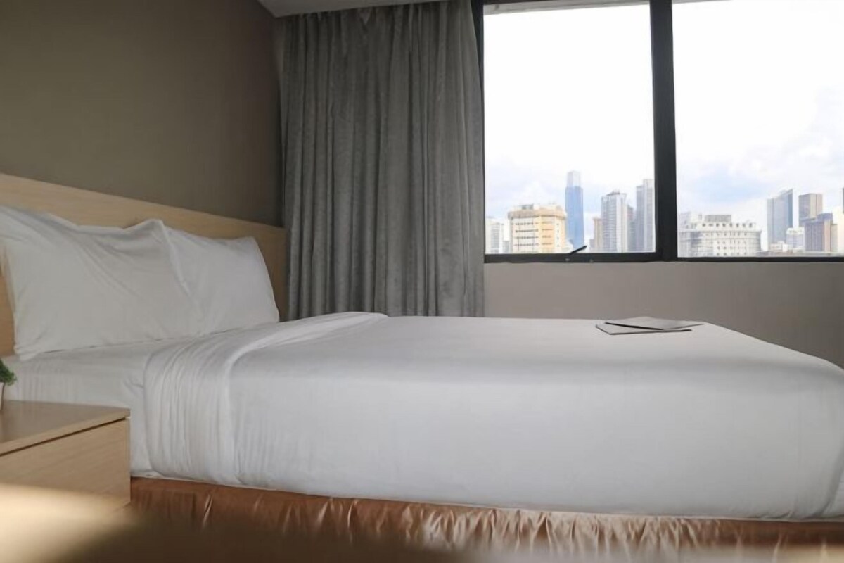 A hotel room with a large bed, a nightstand, a window with a city view, and a curtain.