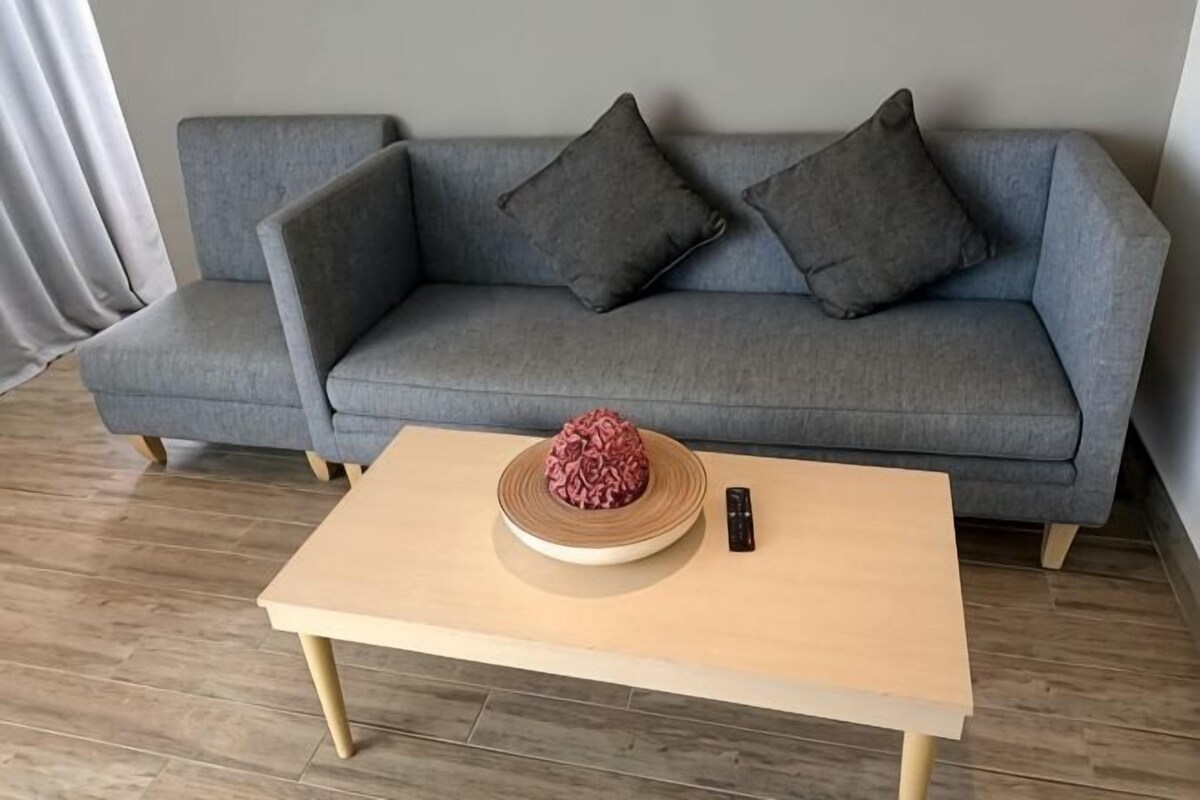 A grey sofa with two cushions, a wooden coffee table with a decorative item and a remote control, and a wooden floor.
