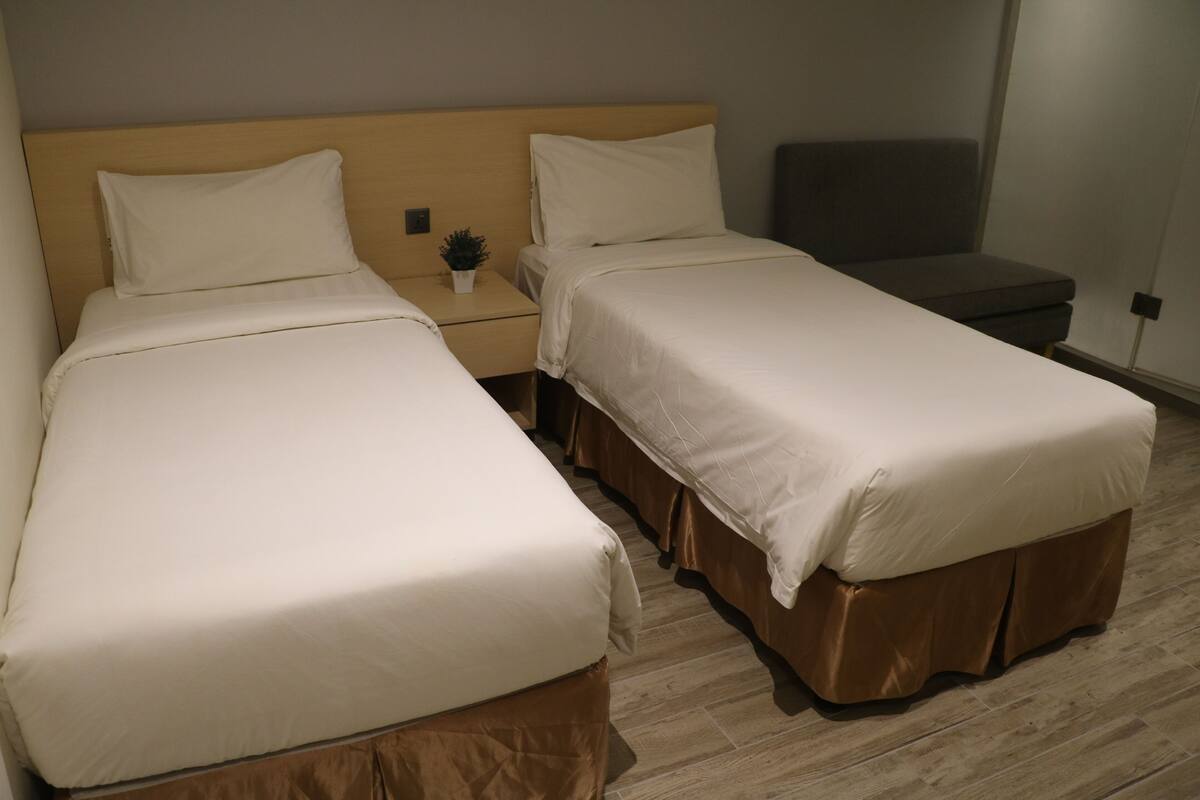Habitación de hotel con dos camas, una mesita de noche con una planta y un banco gris.