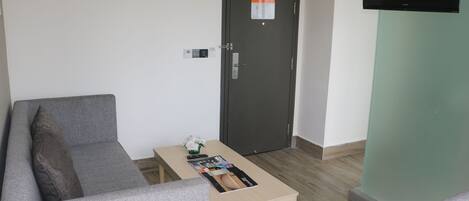 A modern room with a grey sofa, a wooden coffee table with magazines, a door with a notice, and a flat-screen TV on a green wall.