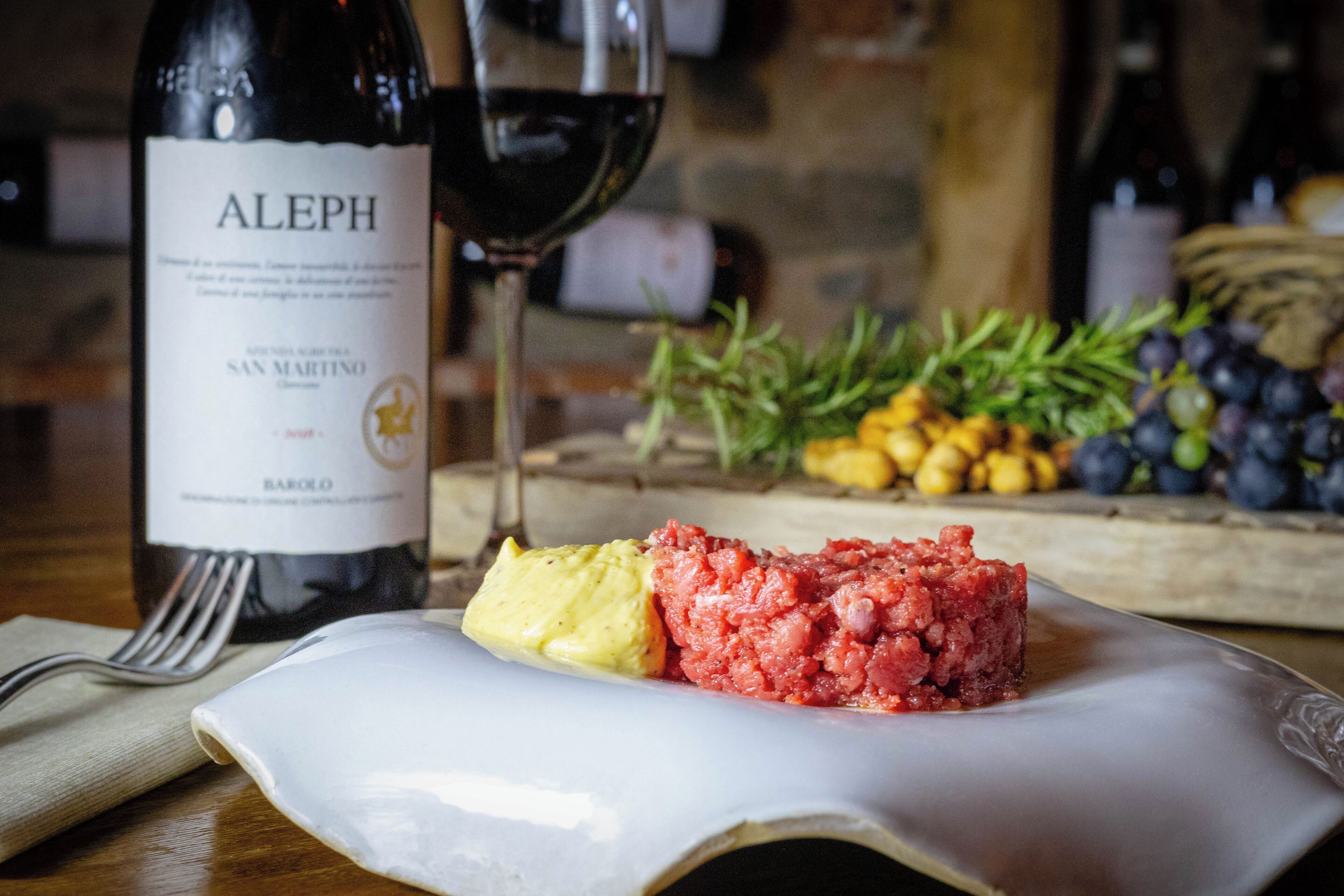 A bottle of Aleph wine, a glass of red wine, and a plate with a food item and some fruits.