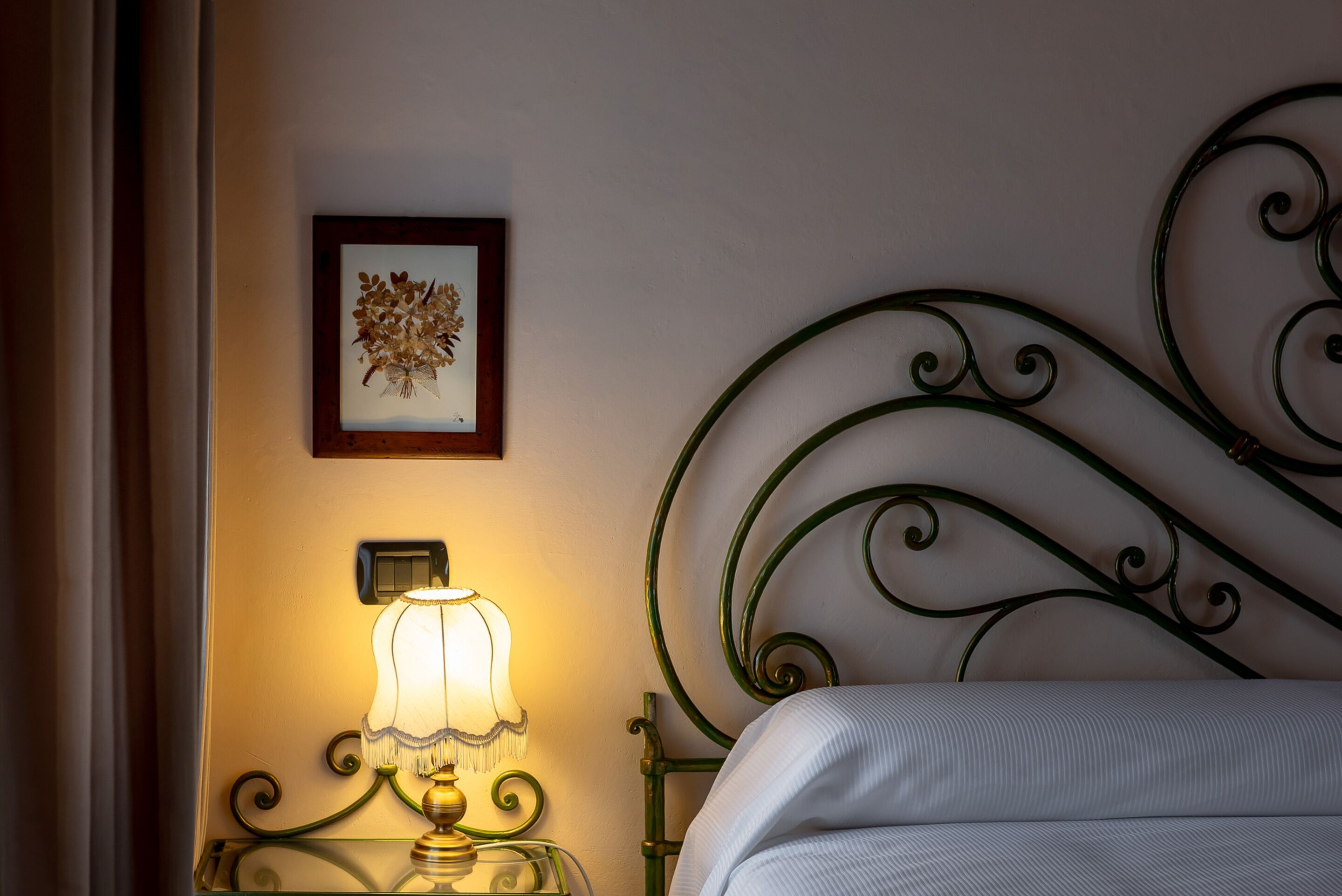 Una cama bien tendida con cabecera metálica verde, una lámpara de noche y un cuadro en la pared.