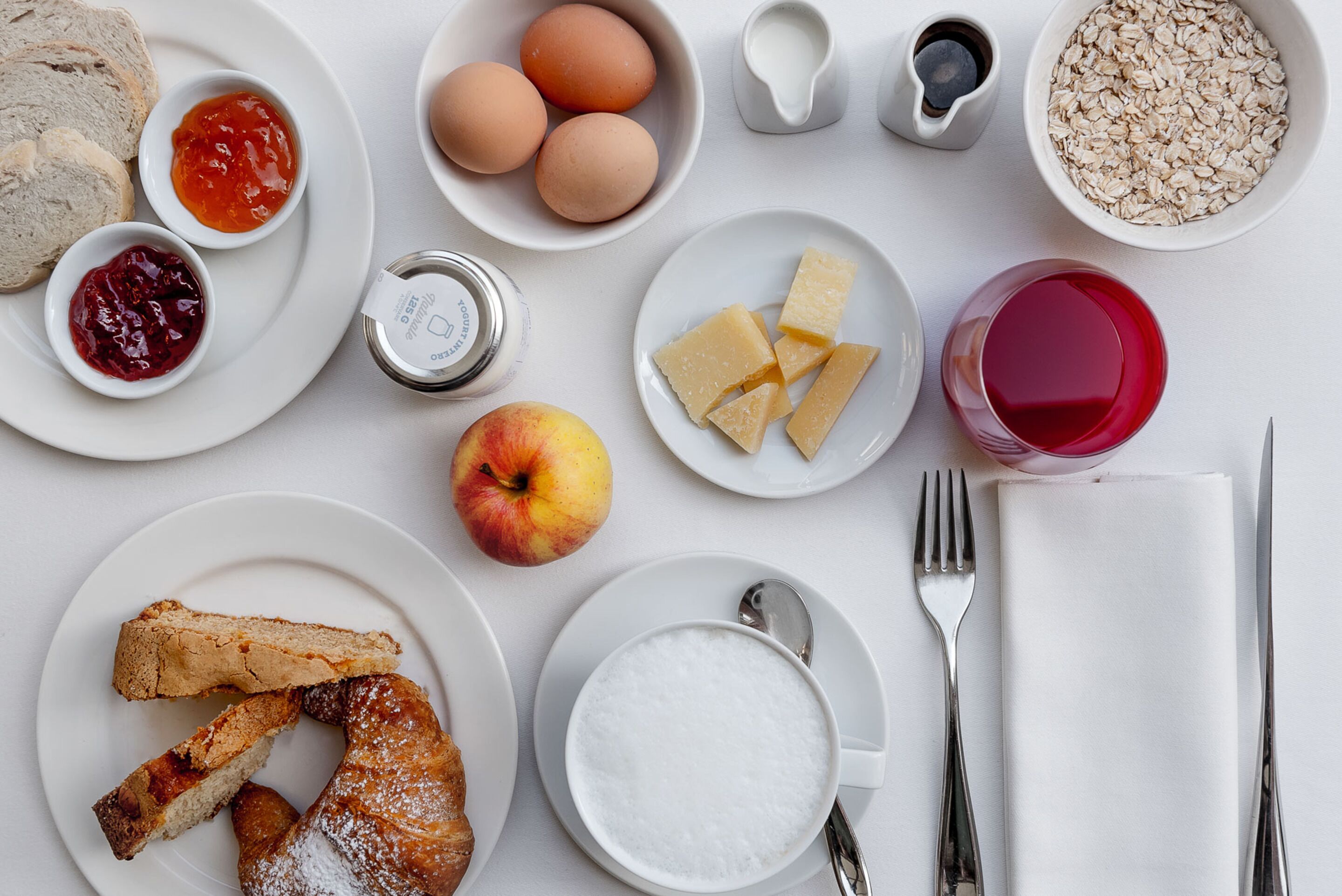 Una mesa de desayuno con pan, huevos, mermelada, queso, avena y una bebida roja.