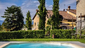 A swimming pool with a clear blue water, surrounded by a well-maintained hedge, and a large house with a tiled roof in the background. - San martino langhe (Clavesana)