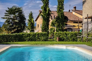 Una piscina con agua cristalina azul, rodeada por un seto cuidado y una casa grande con techo de tejas al fondo.