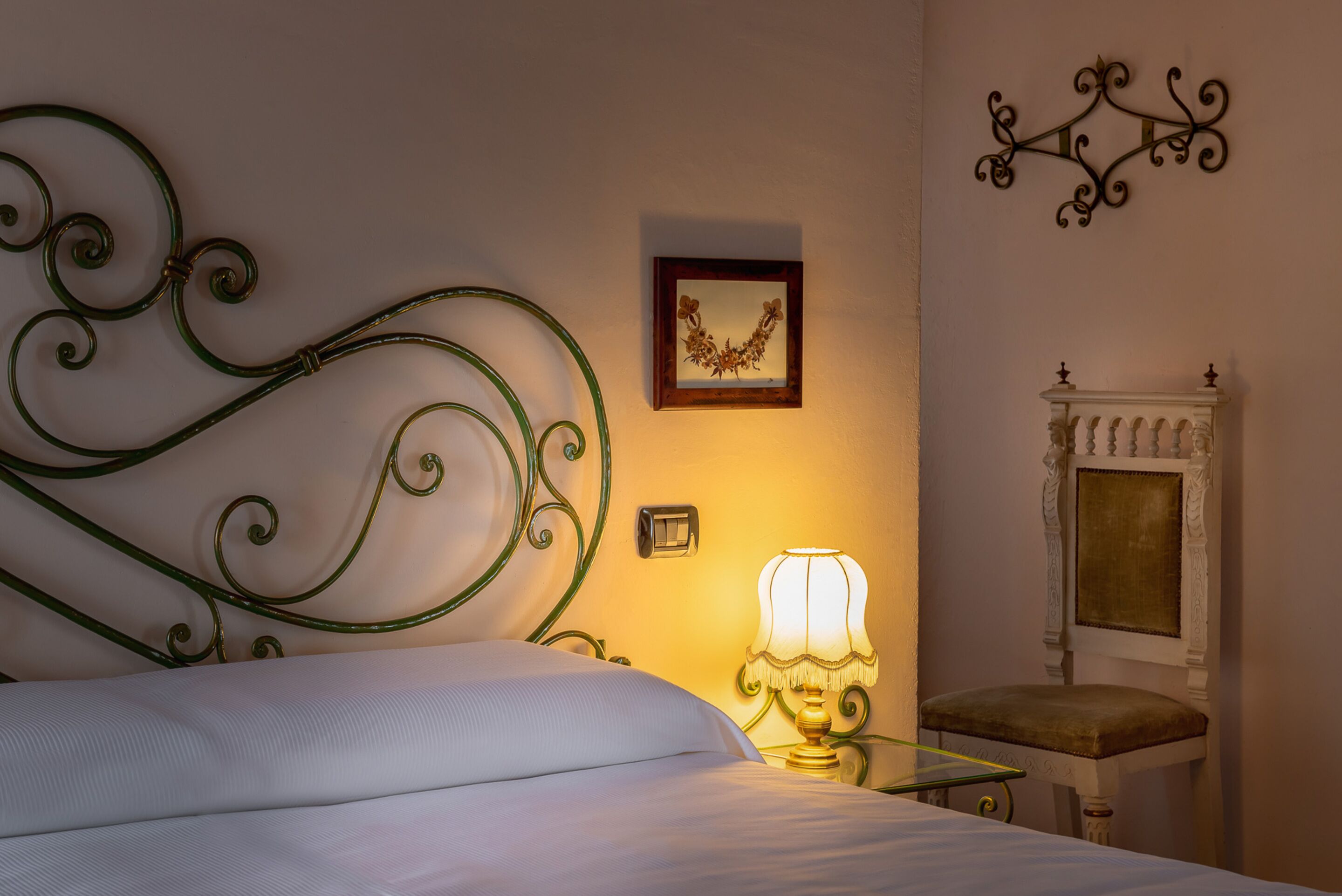 A neatly made bed with a green metal headboard, a bedside lamp, a framed picture on the wall, and a small white table with a chair.