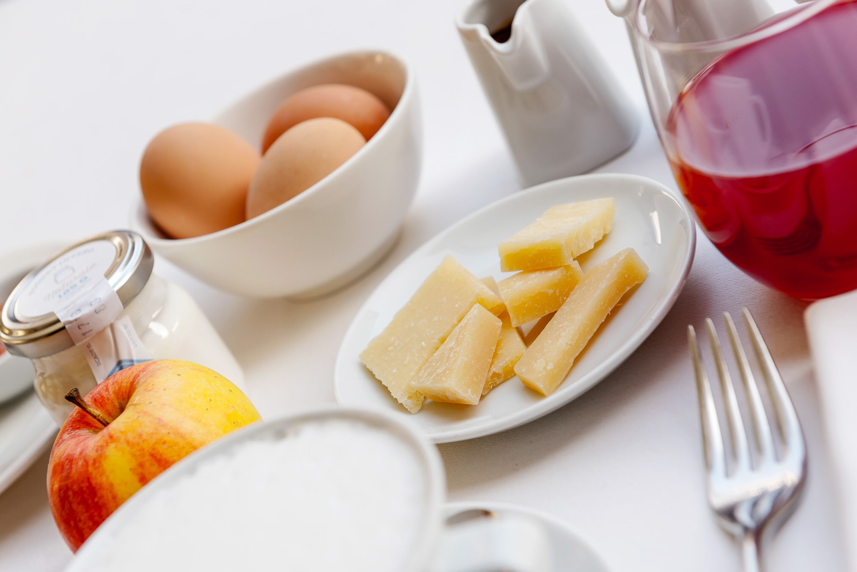 Desayuno con huevos, queso, una jarras de líquido rojo, un frasco y un tenedor sobre una mesa blanca.